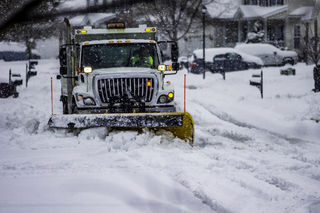 Snowplow service truck clearing residential roads of snow while flakes are still falling